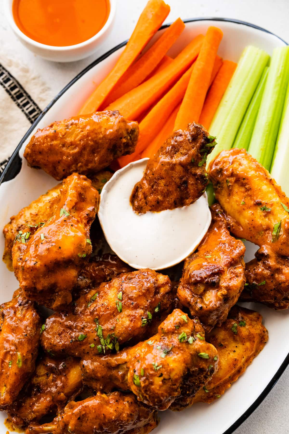 A large plate of air fried chicken wings with one wing in a small container of ranch. There's a side of celery and carrot sticks.