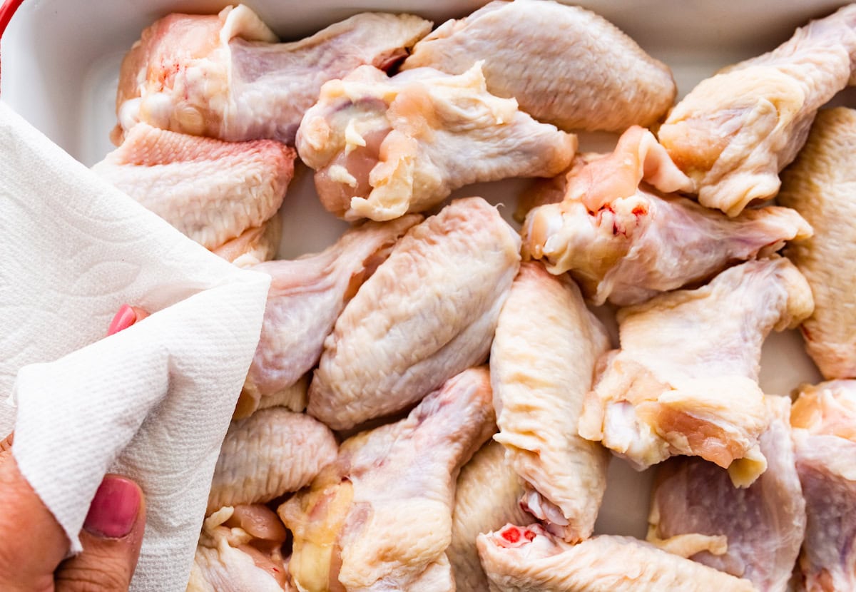Multiple raw chicken wings in a container are being patted down dry with a paper towel.