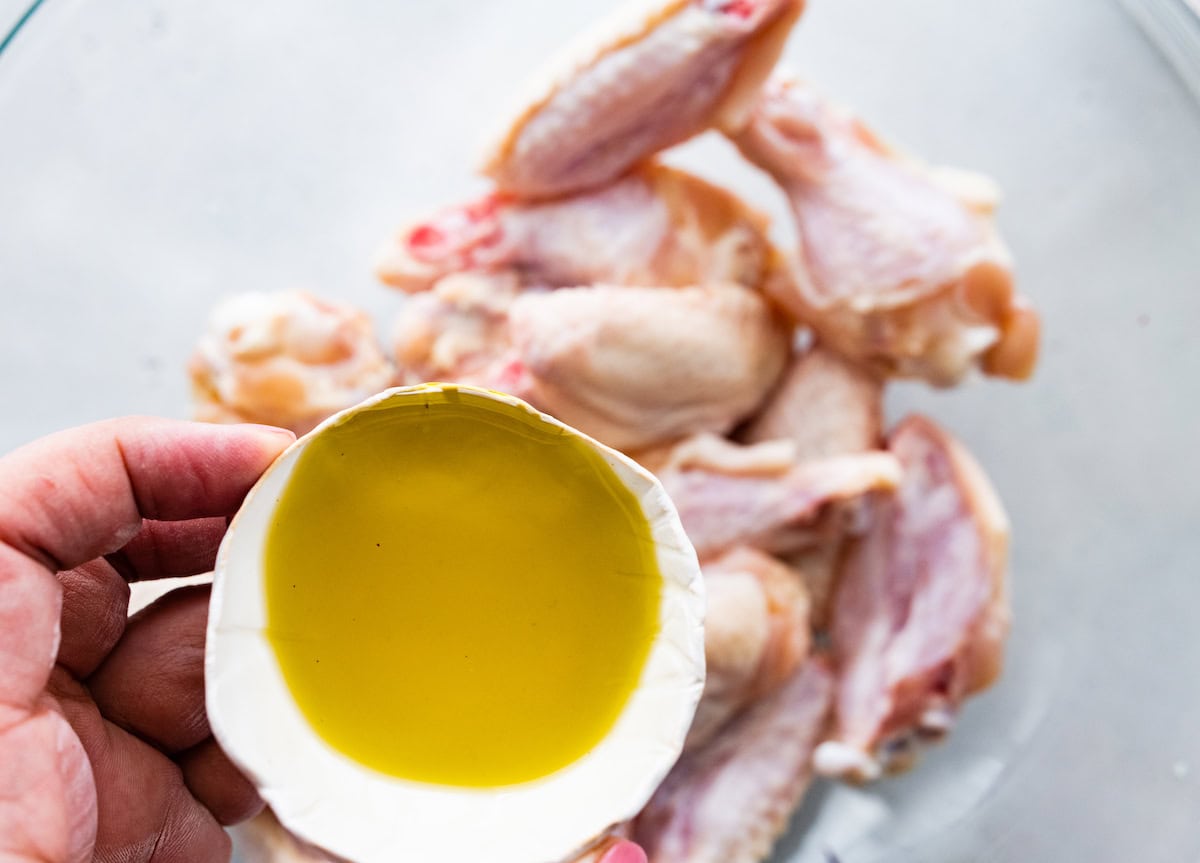 A hand holds a small container of olive oil over a bowl of raw chicken wings.