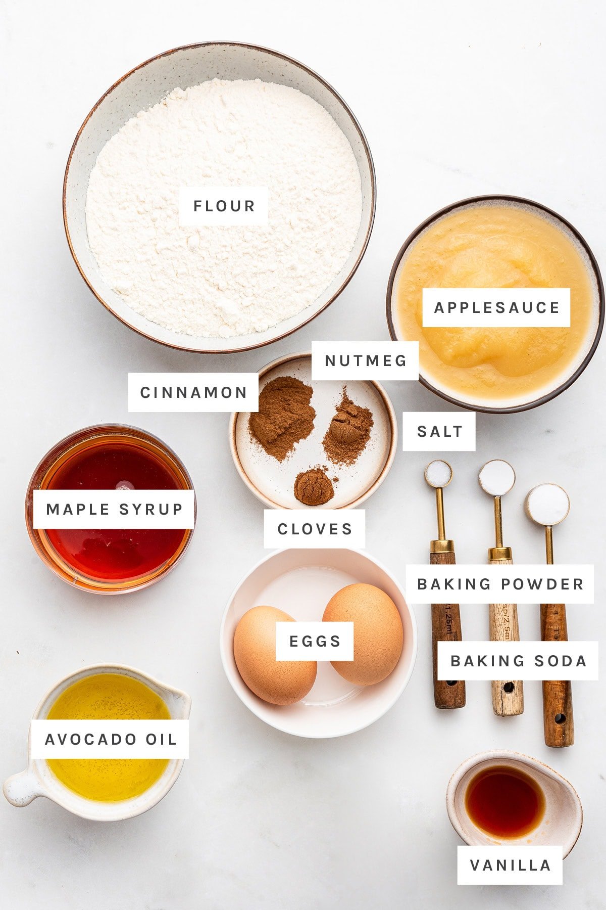 Ingredients measured out to make Applesauce Cake: flour, applesauce, nutmeg, cinnamon, cloves, salt, maple syrup, baking powder, baking soda, eggs, avocado oil and vanilla.
