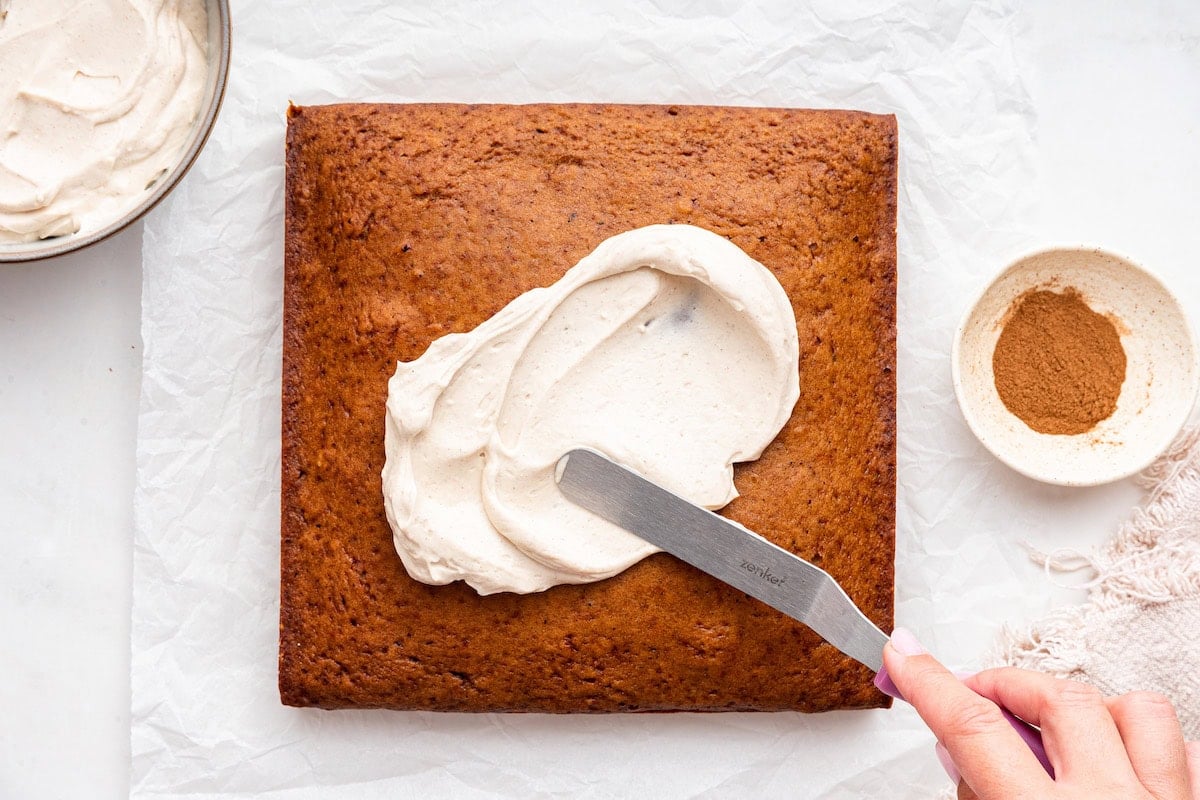 A womans hand uses a spatula to spread a cream cheese frosting on an applesauce cake.