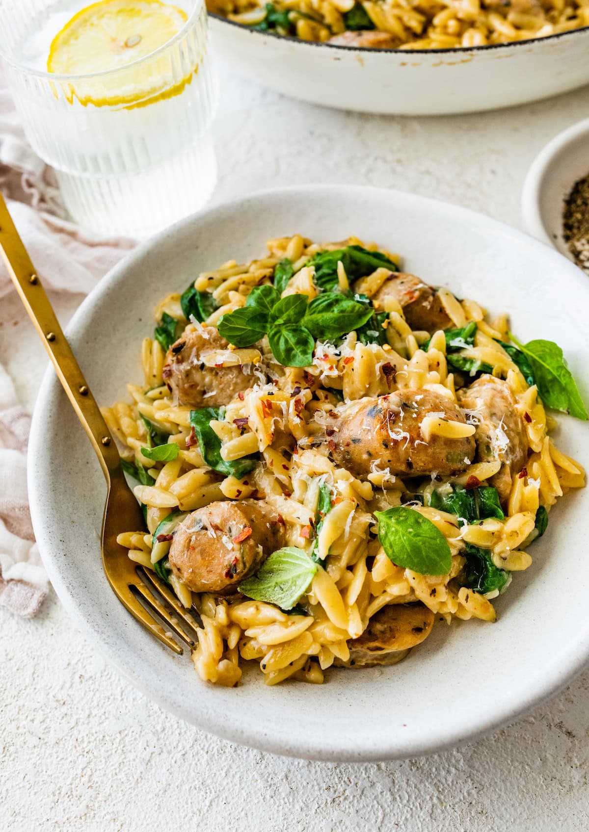 A white shallow bowl with orzo, chicken sausage and spinach with a gold fork laying on the left hand side.