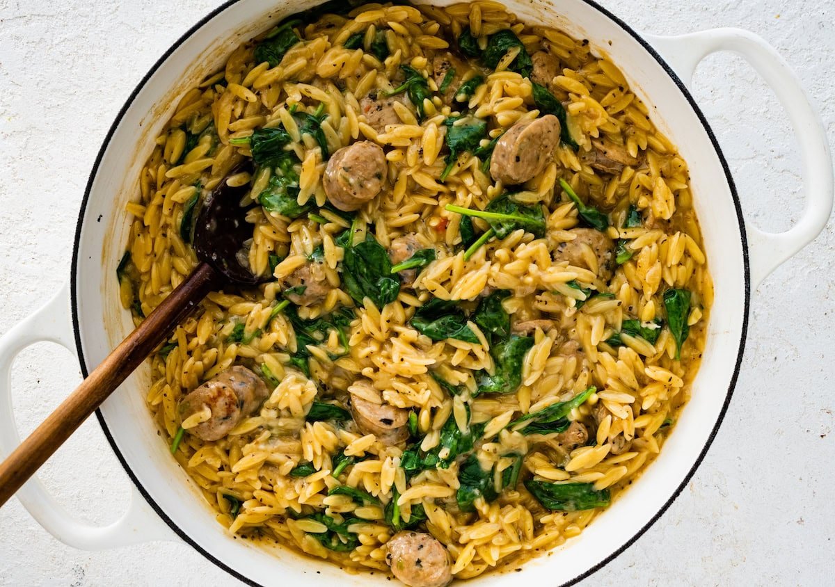 A white Dutch oven with cooked orzo, chicken sausage and baby spinach.