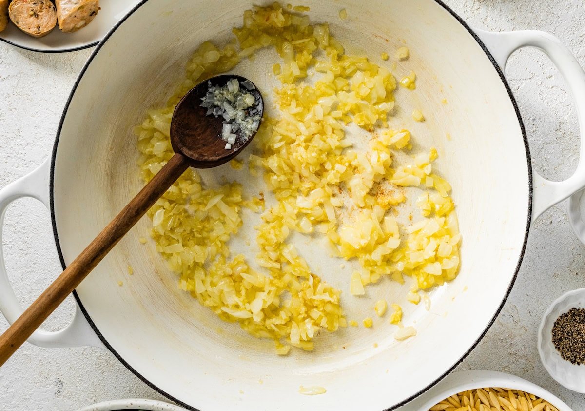A white Dutch oven with oil cooked chopped onions and minced garlic as well as a wooden spoon.