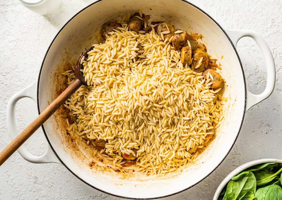 A white Dutch oven with cooked chicken sausage and dry orzo on top.