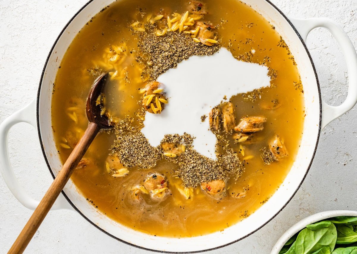 A white Dutch oven with orzo, broth, chicken sausage, coconut milk and pepper.