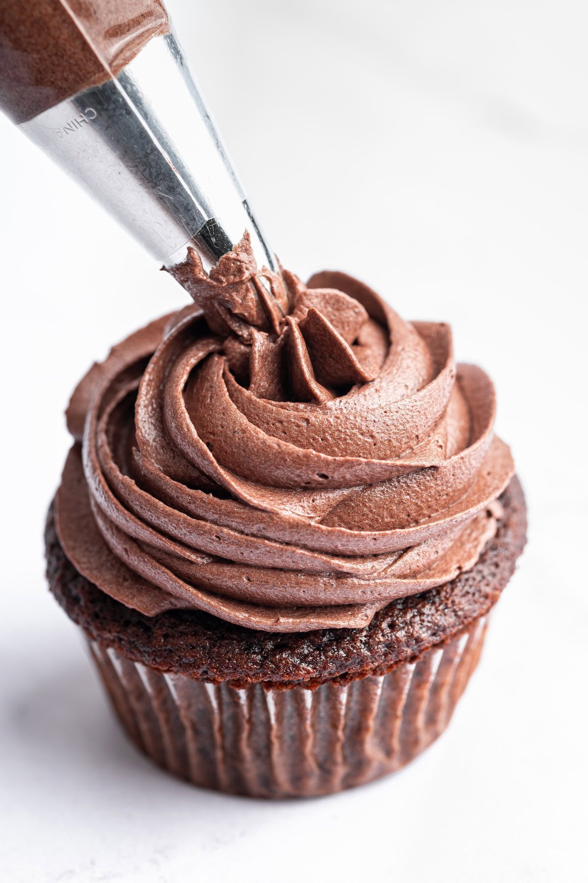 Chocolate buttercream frosting being piped onto a chocolate cupcake from a piping bag.