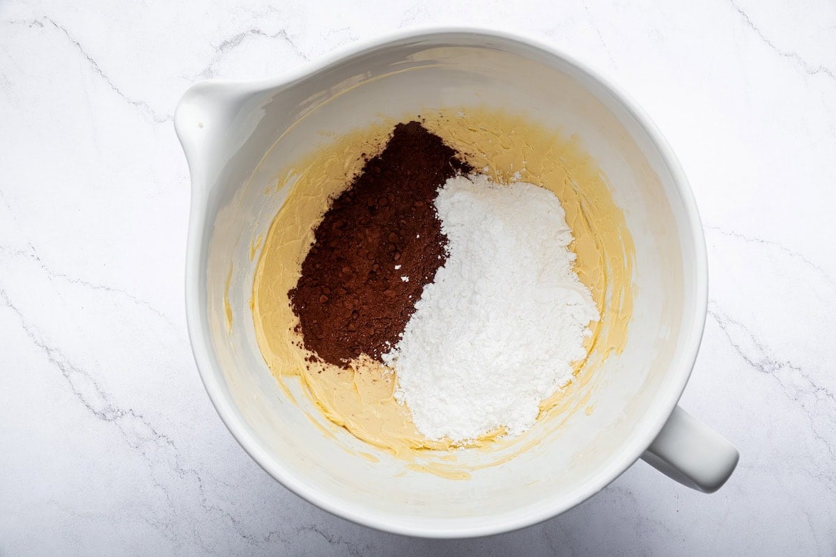 Powdered sugar and cocoa powder added to whipped butter in a mixing bowl.