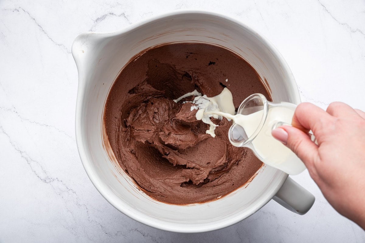 A woman's hand pouring milk into a mixing bowl with the whipped chocolate buttercream frosting.