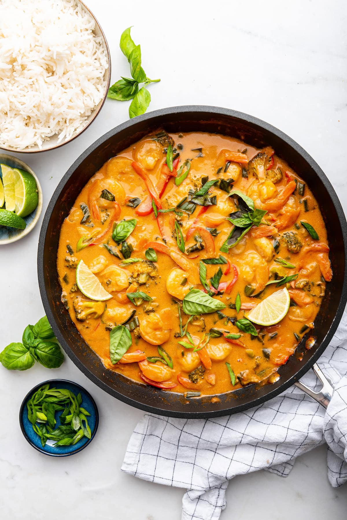 A large skillet with coconut shrimp curry. There is a bowl of white rice and some lemon wedges on the side.