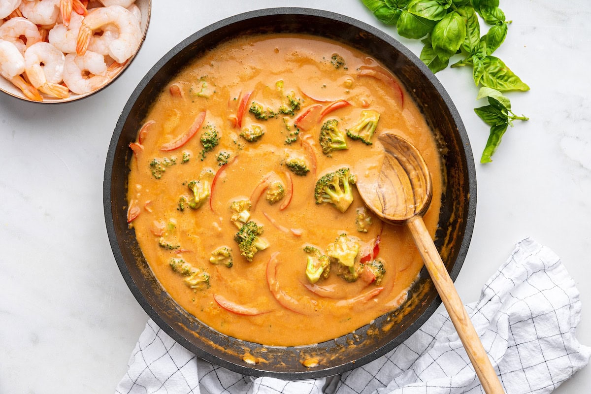 A skillet with broccoli, coconut milk, curry paste, and red bell peppers.