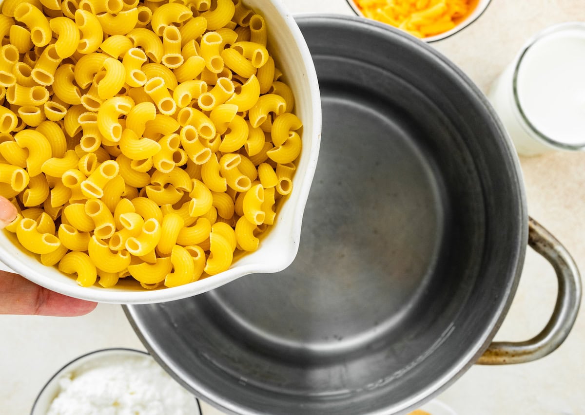 Raw macaroni noodles in a bowl are being added to a large pot of water.