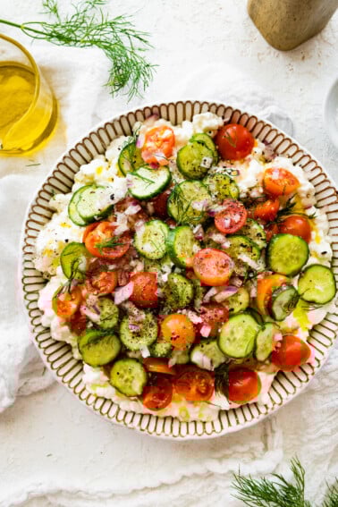 A cottage cheese salad with cucumber, tomatoes, seasoning, and fresh herbs in a large bowl.