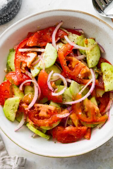 In a small white bowl, a cucumber tomato salad with sliced red onion, salt, and pepper.
