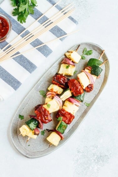 Grilled BBQ tempeh and veggie skewers on an oval platter, next to skewers on a towel.