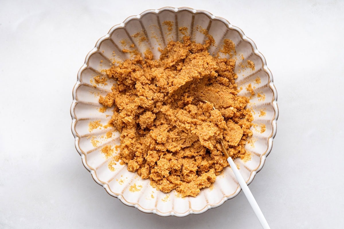 Graham cracker crumbs and maple syrup mixed together in a small mixing bowl with a white spoon.