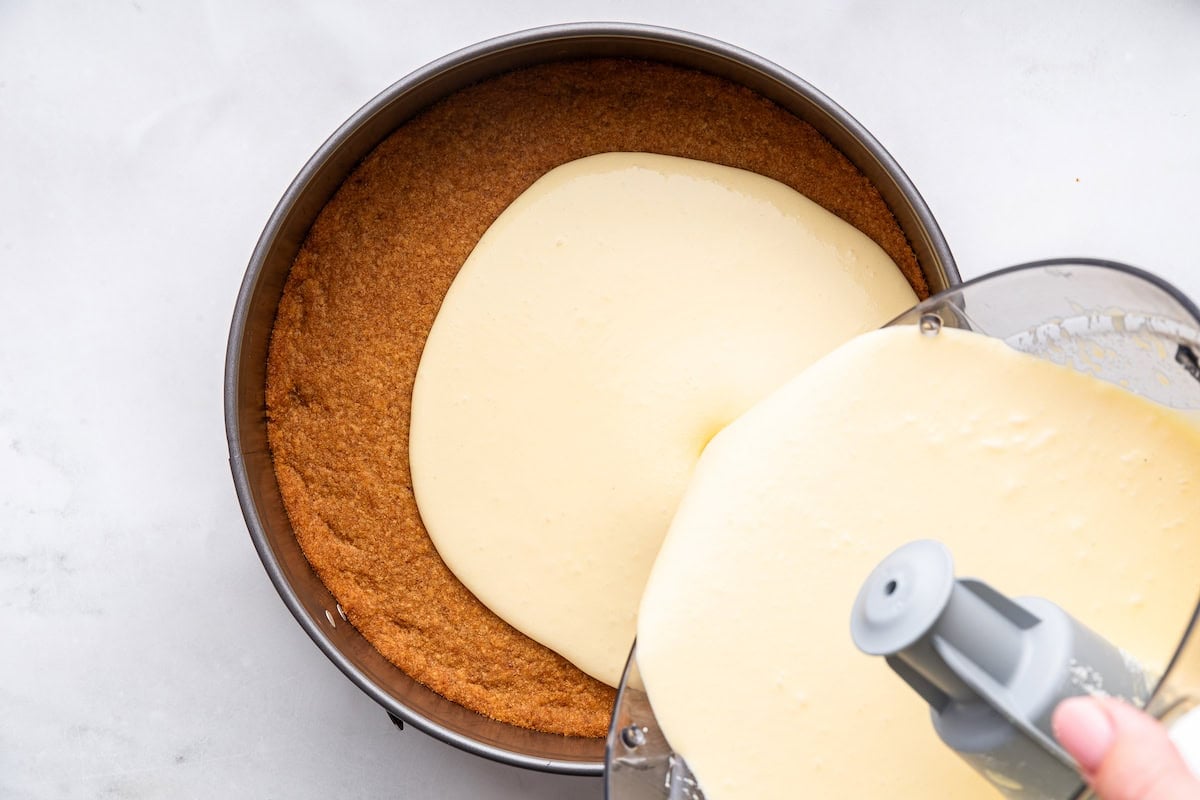 The cheesecake filling being poured onto the cheesecake crust from the bowl of a food processor.