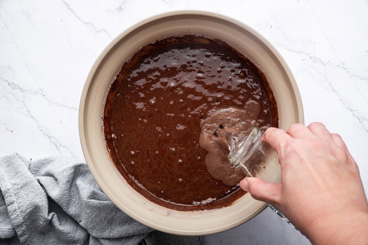 Boiled water is being poured into a large mixing bowl of chocolate cake batter.