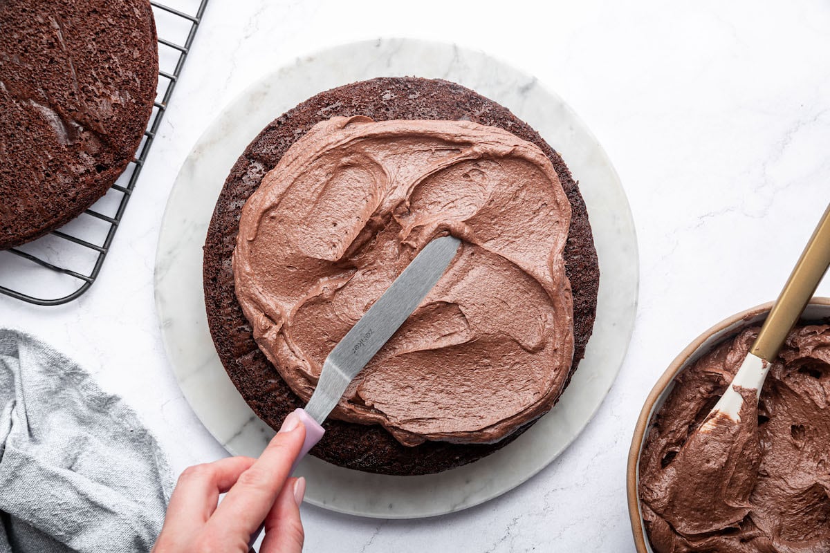 A chocolate buttercream frosting is spread on a chocolate cake using a metal spatula.