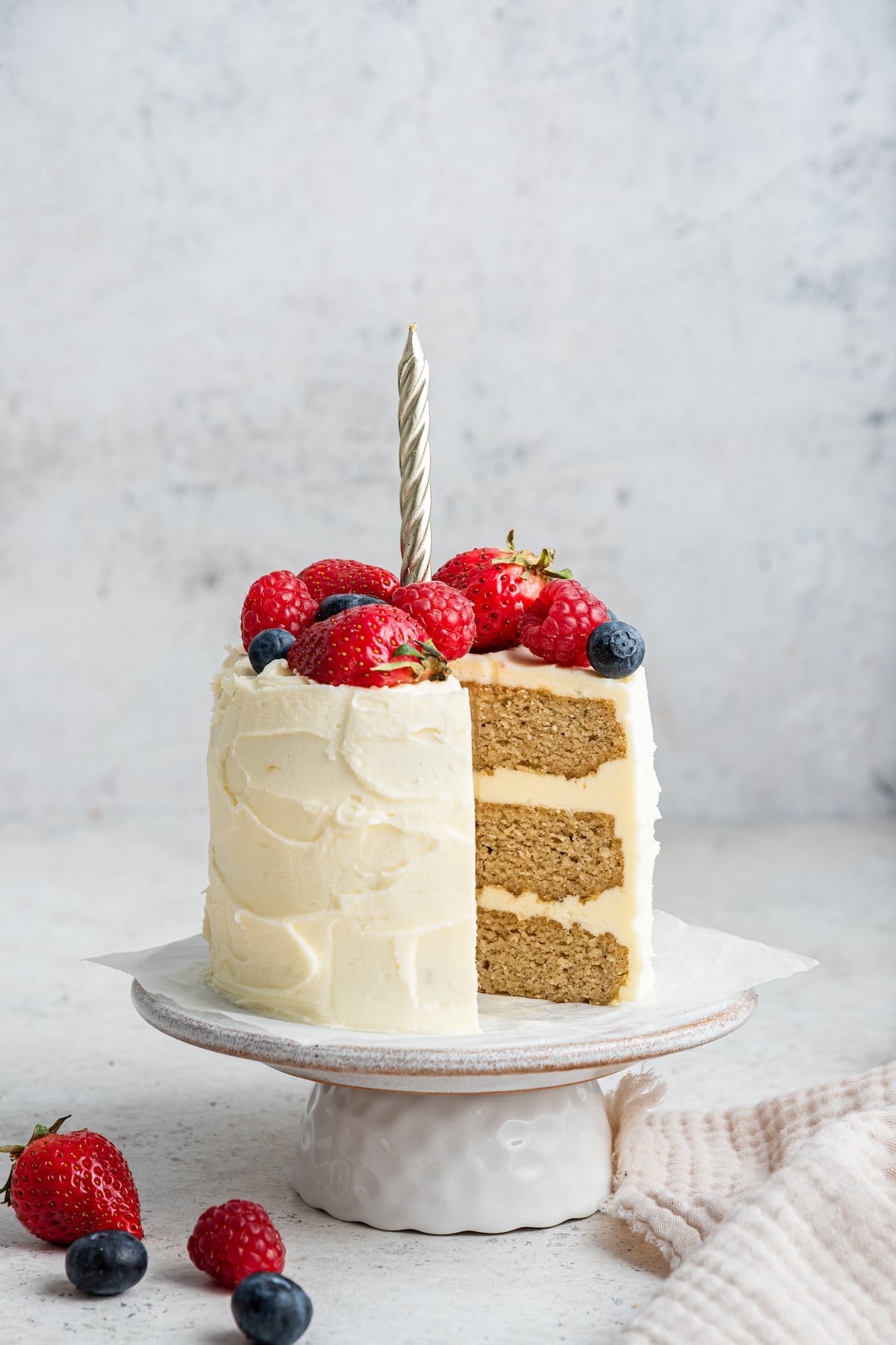 A frosted smash cake with a candle topped with fresh berries sitting on a cake stand. One slice of the cake has been cut out of it.
