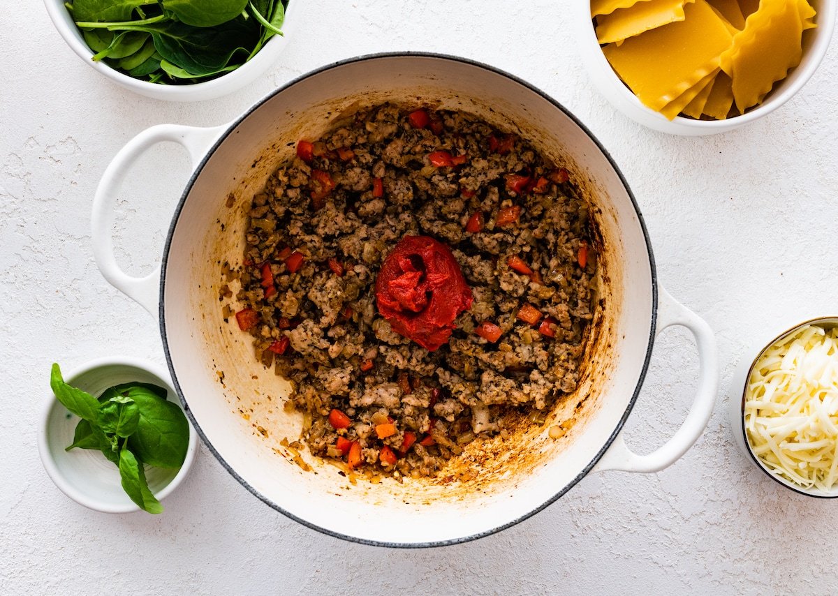 Tomato paste added to a large pot of cooked ground sausage, spices, onions, red bell pepper, and garlic.