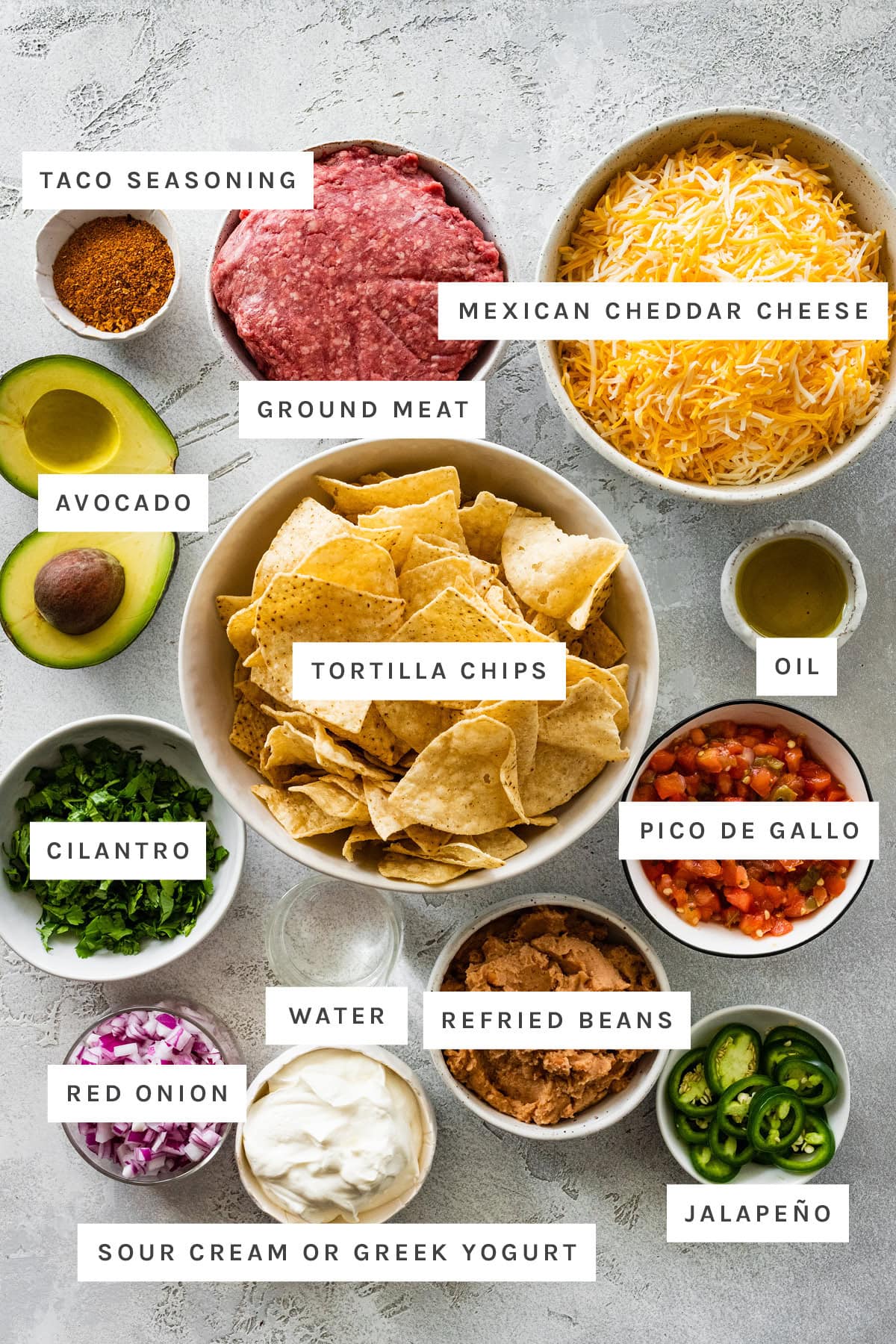 Ingredients measured out to make Loaded Nachos: taco seasoning, ground meat, Mexican cheddar cheese, avocado, tortilla chips, oil, pico de gallo, cilantro, water, refried beans, red onion, sour cream and jalapeño.
