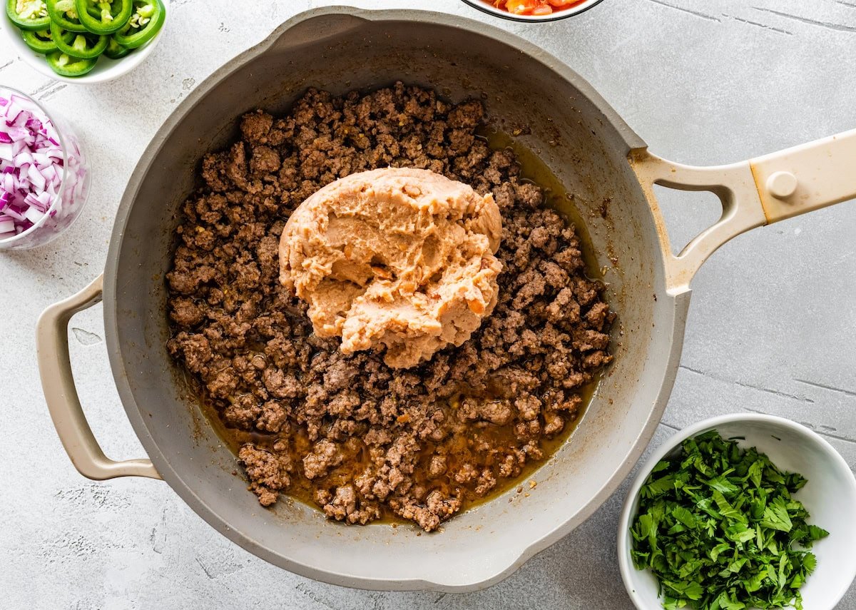 Refried beans added to a pan of cooked ground beef.