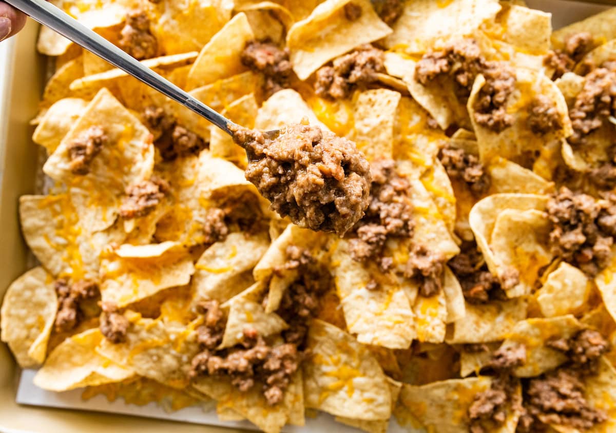 A spoon holding a portion of meat and beans over a tray of tortilla chips.