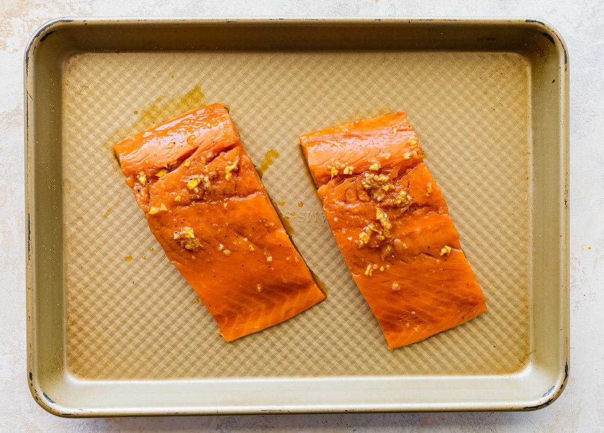 Two marinated salmon filets on a small baking tray before being baked.