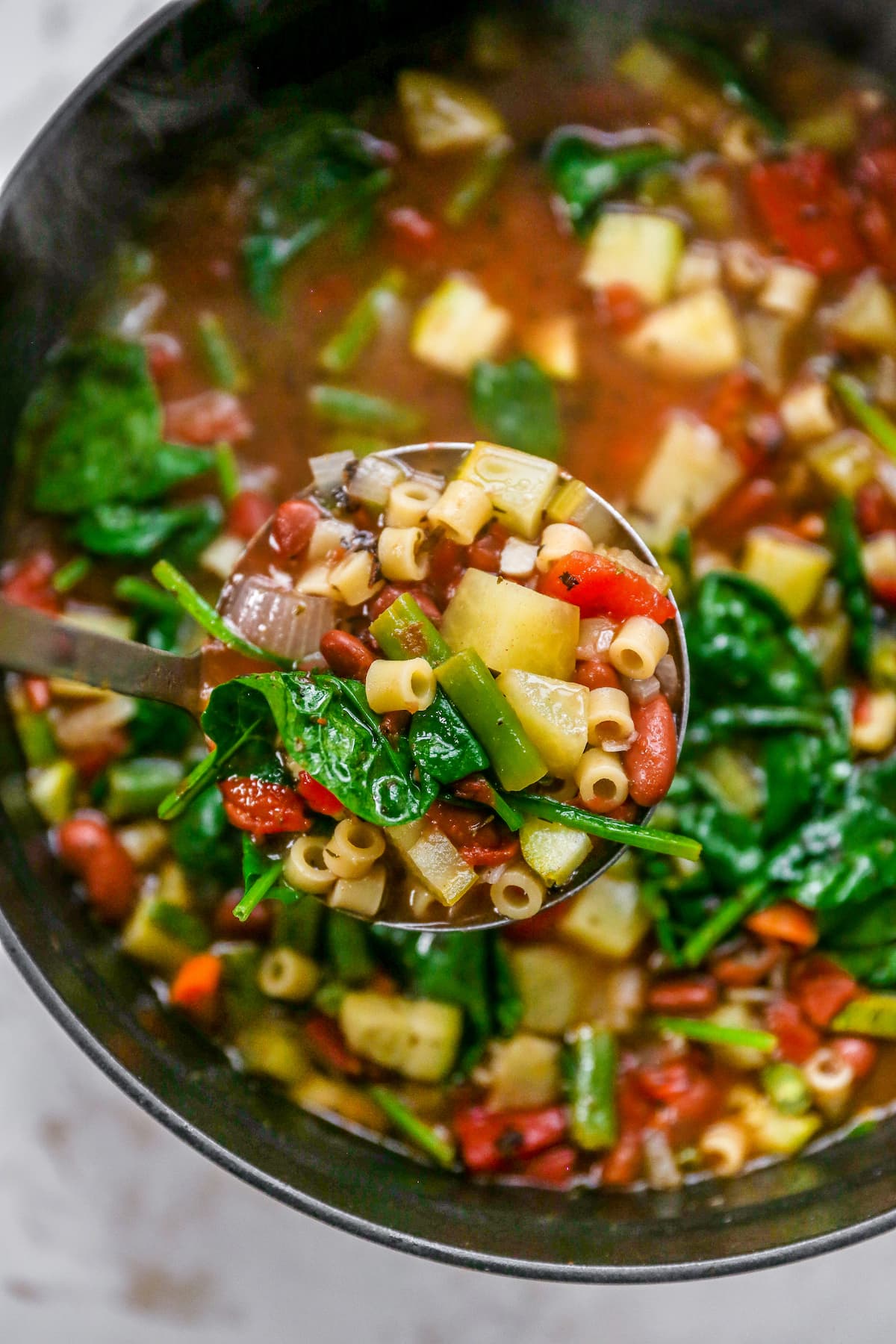 An overhead photo looking down into a large pot of minestrone soup. A ladle is holding up a scoop of the soup close to the lense.