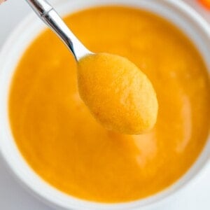 Up close shot of peach puree on a small sliver spoon hovering over a white bowl with peach puree.