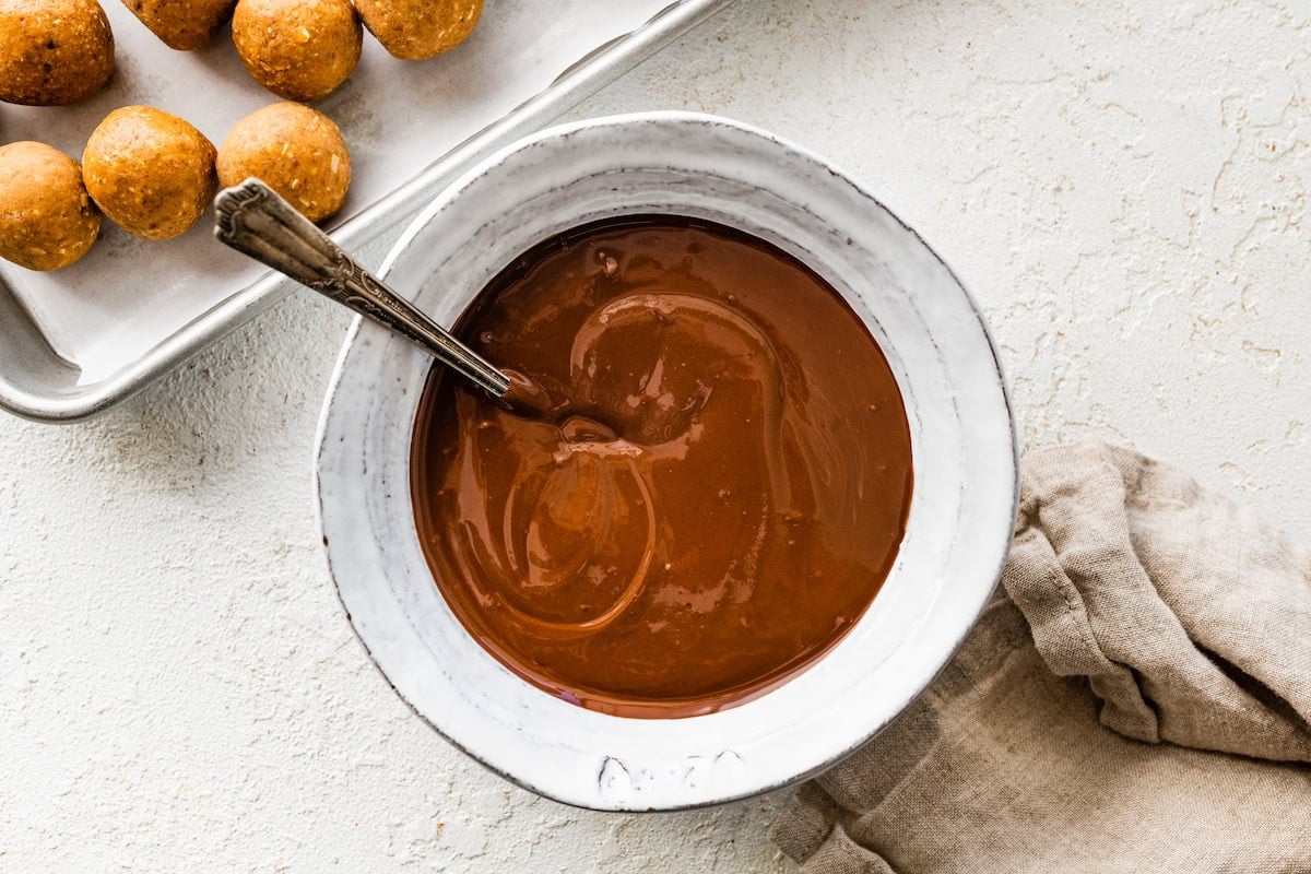 Melted chocolate in a bowl with a spoon near a tray of peanut butter balls.