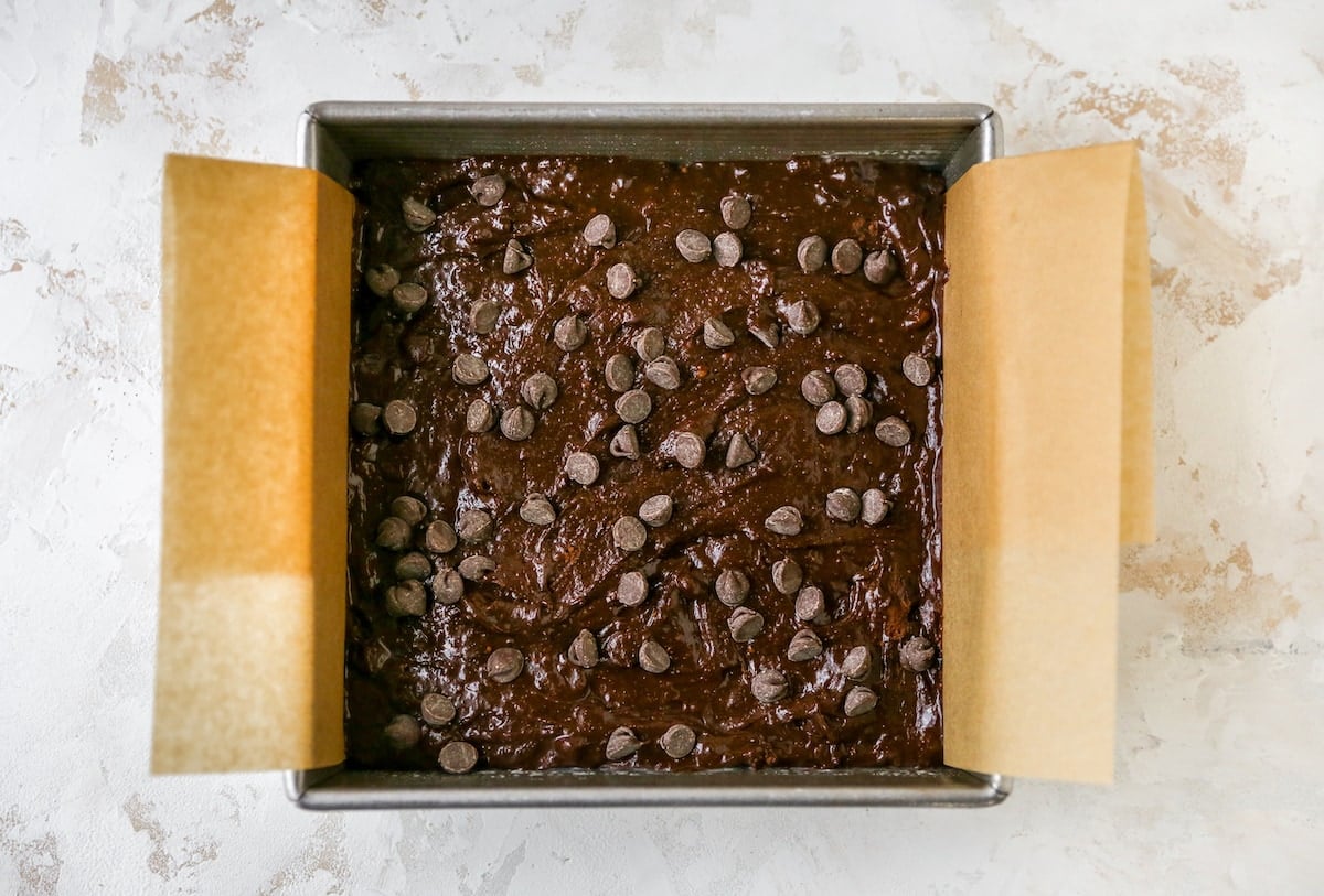 An 8x8 baking pan lined with brown parchment paper. The brownie batter has been poured into the pan and it has been topped with chocolate chips.