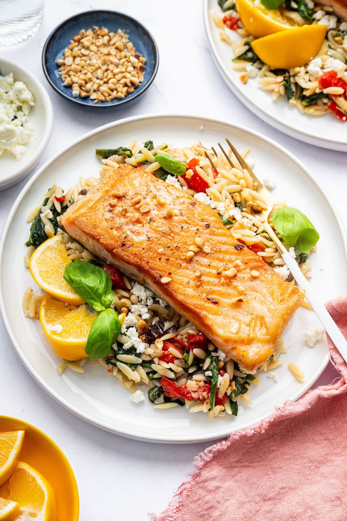 A salmon filet served on a white plate over a bed of orzo combined with roasted red peppers, chopped dates, feta and swiss chard. A fork is on the plate next to the salmon.