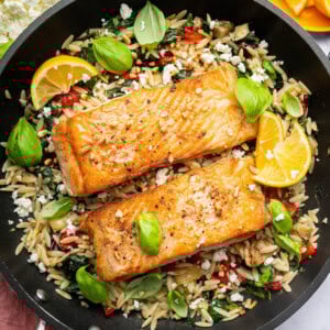 Two salmon filets in a skillet served over a bed of orzo combined with roasted red peppers, chopped dates, feta and swiss chard. It's topped with lemon slices and fresh basil.