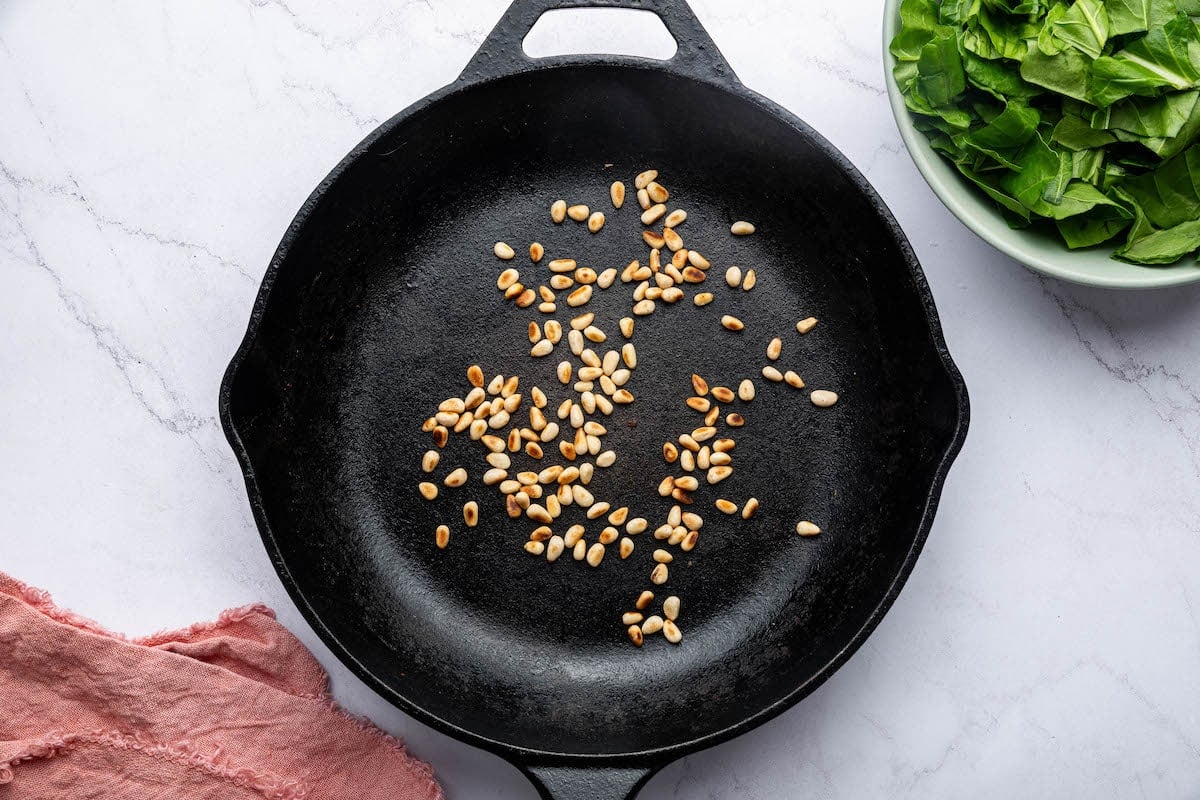 Toasted pine nuts in a cast iron skillet next to a bowl of chopped swiss chard.