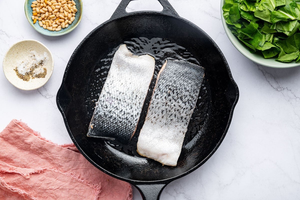 Two salmon filets cooking in a cast iron skillet, skin side up. A bowl of pine nuts and chopped swiss chard is next to the skillet.