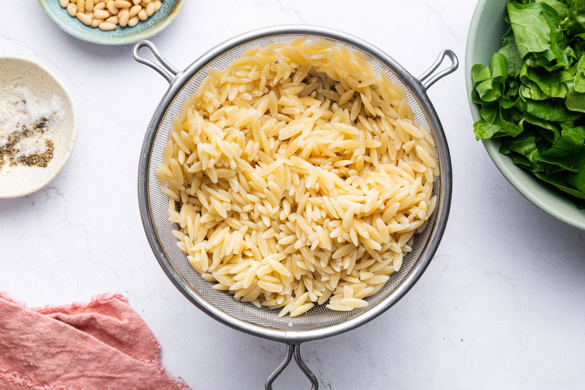 Cooked orzo in a strainer next to a bowl of chopped swiss chard and pine nuts.