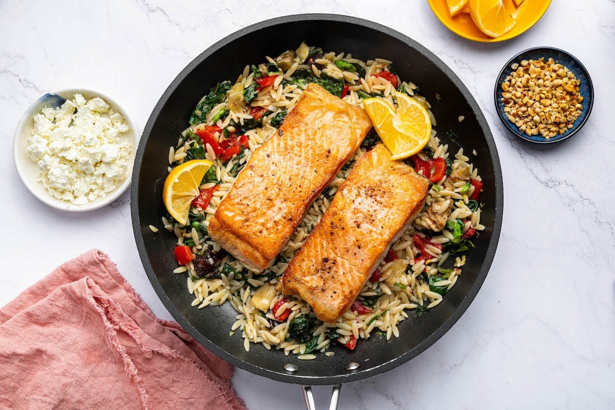Two salmon filets in a skillet served over a bed of orzo combined with roasted red peppers, chopped dates, feta and swiss chard. It's topped with lemon slices and fresh basil.