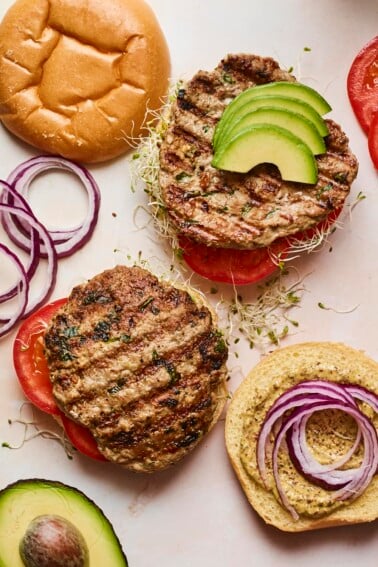 Two open faced spinach and feta turkey burgers with burger toppings and buns around on the table.