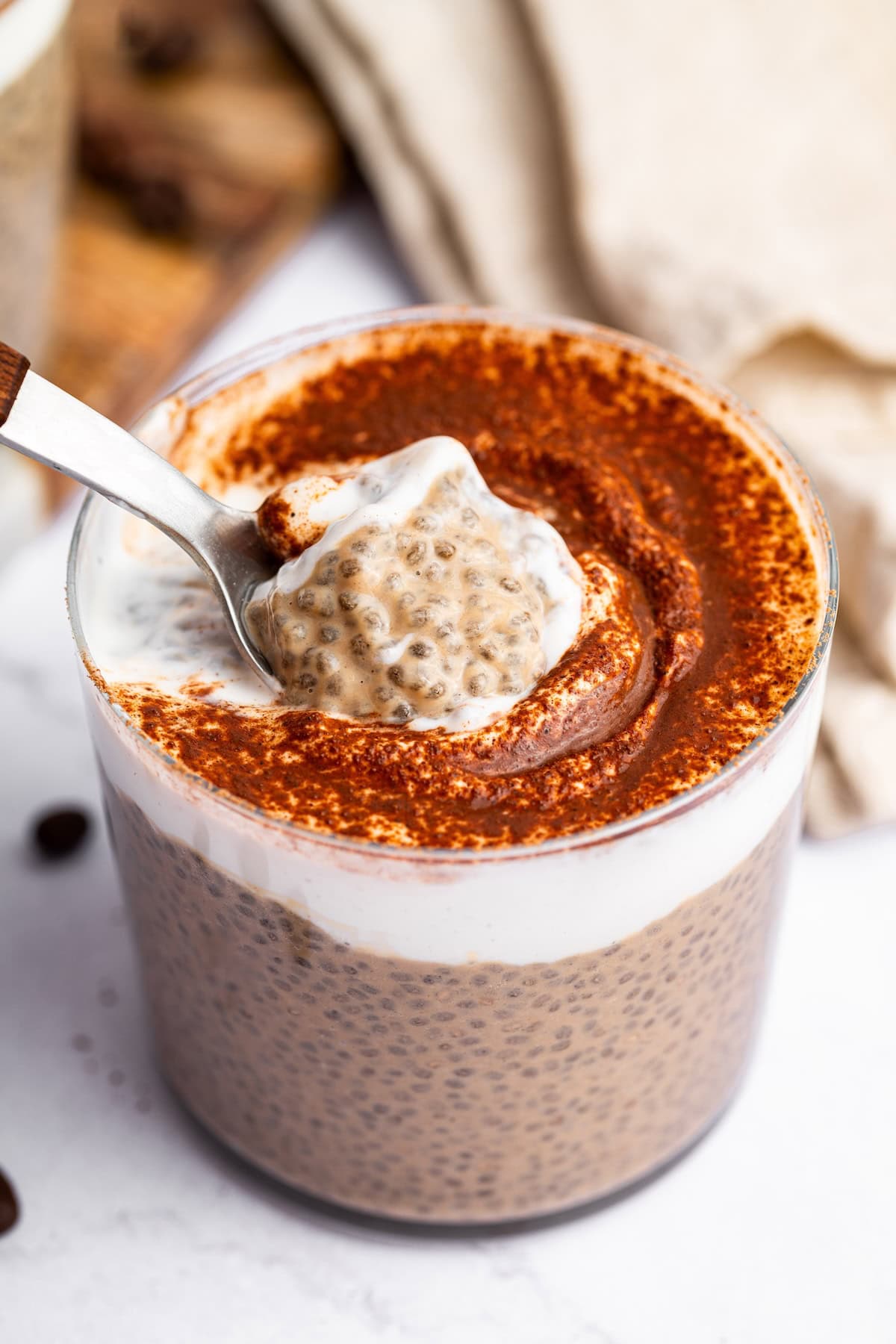 A spoon digging in to a glass cup of tiramisu chia pudding that is topped with cocoa powder.