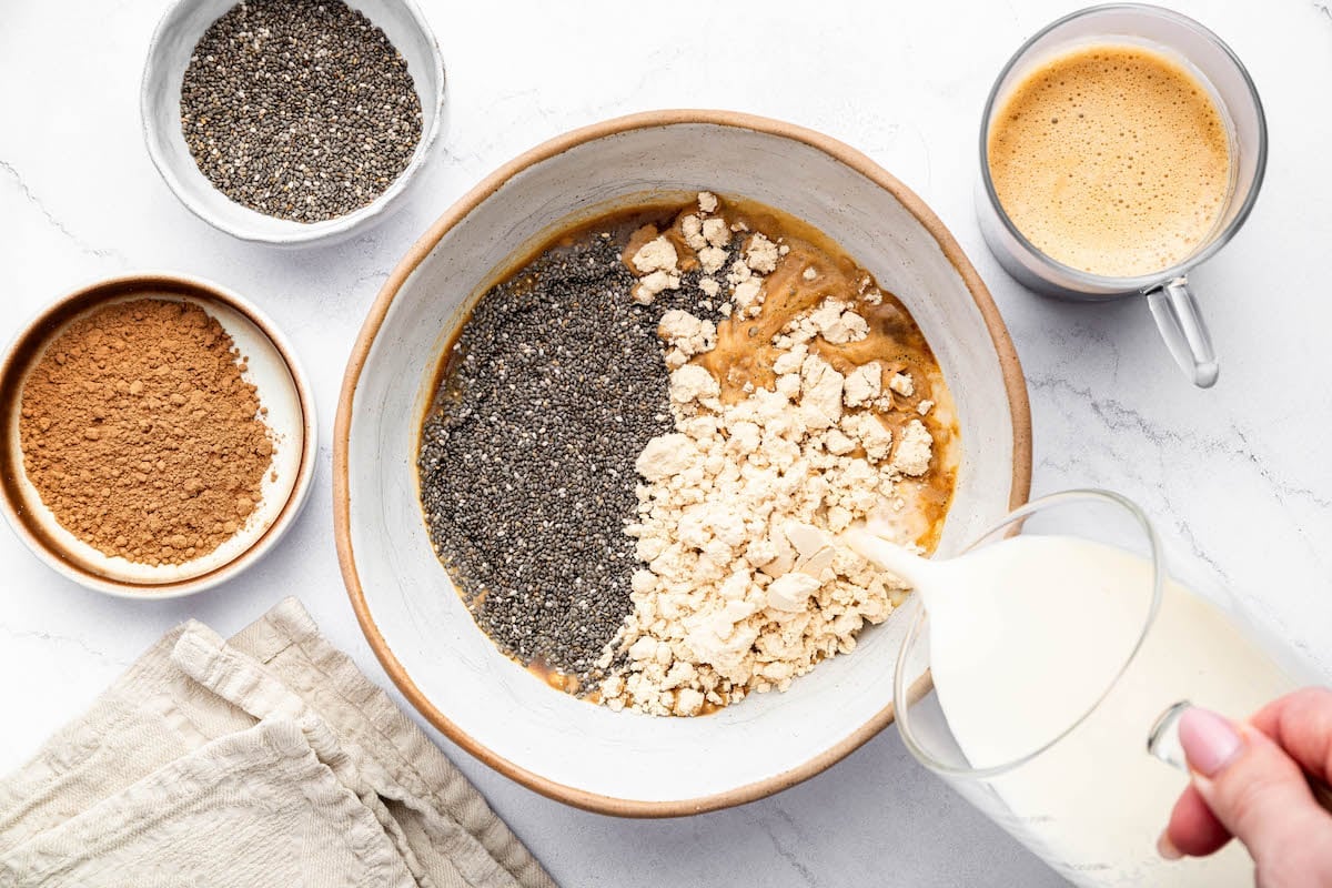 Almond milk being poured into a bowl of chia seeds, maple syrup, protein powder and espresso.