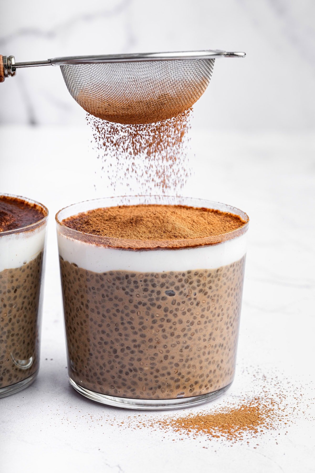Two glass cups of tiramisu chia pudding near one another. A small metal sifter is being used to add cocoa powder on top of the pudding.