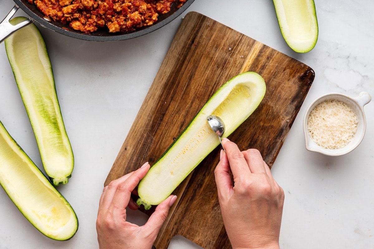 A womans hand uses a small spoon to scoop out the flesh of a zucchini to create a "boat".