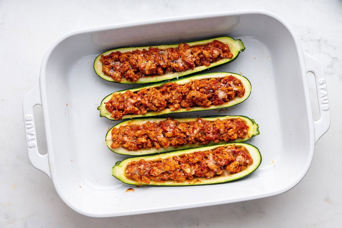 Four zucchini boats stuffed with seasoned turkey topped with fresh cheese in a large baking dish.